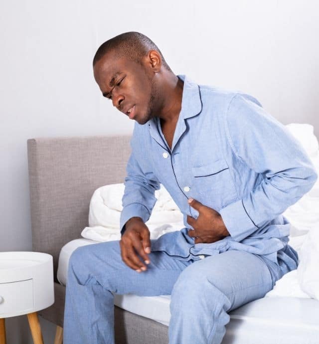 man in blue pajamas sitting on his bed holding stomach in pain. He has abdominal pain.