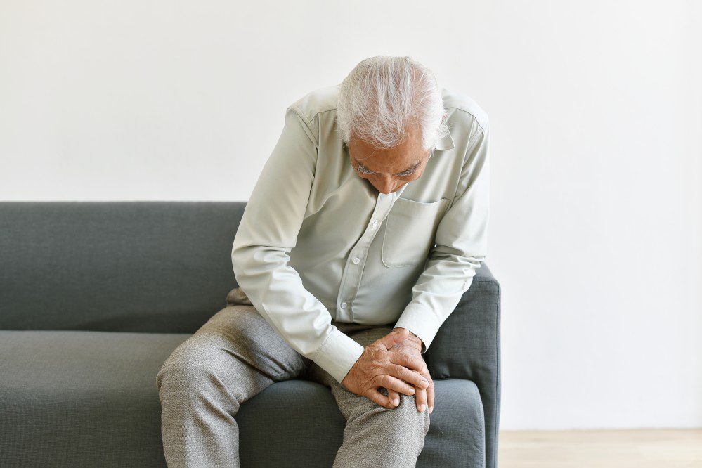 old man dressed in olive pit colour t-shirt and grey pants seated on a couch holding knee in pain. He is at a chronic pain specialist office.
