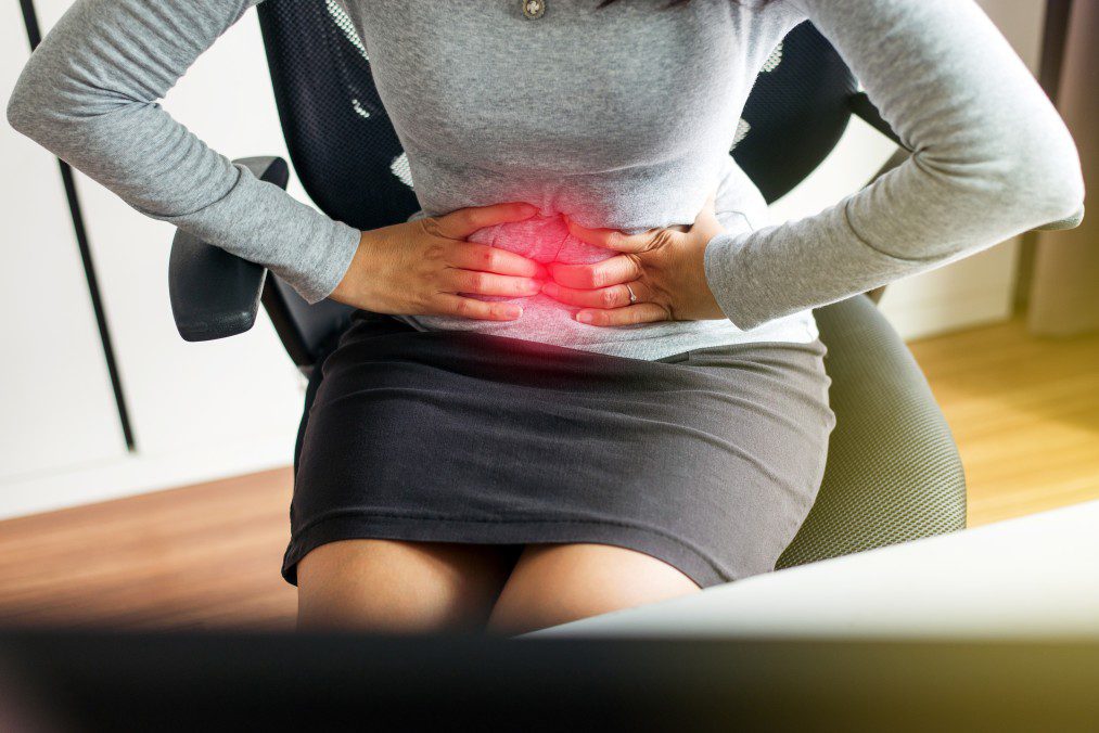 person wearing a grey long-sleeve shirt and black skirt holding her stomach in pain with the affected region highlighted in red. She is experiencing pelvic pain.