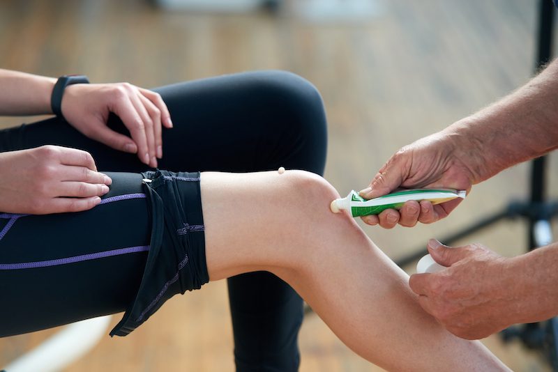 Therapist applying topical agent ointment cream on female knee.