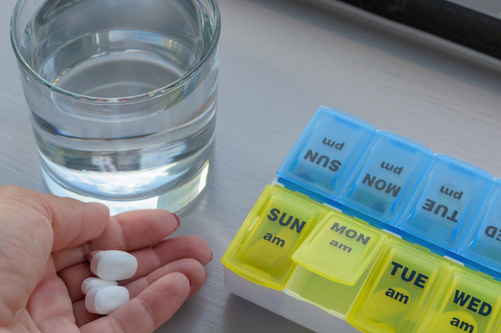 person holding white tablets with hand placed on a grey desk next to a glass containing water and blue and yellow pill planner